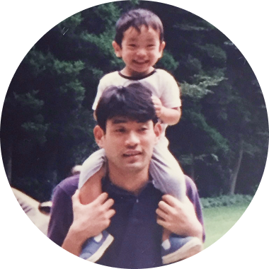 幼い男の子が父親の肩に乗り、緑豊かな屋外で笑顔を見せている温かみのある親子の写真