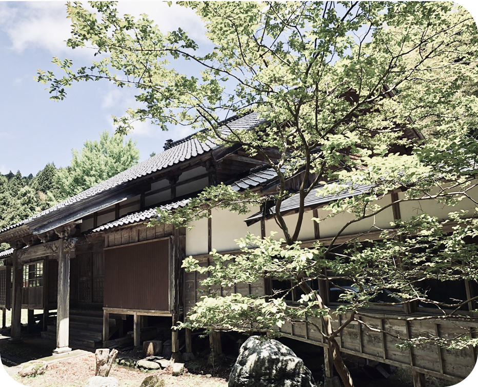 木造の日本家屋が緑の木々に囲まれ、縁側と庭が調和した静かな田舎の風景写真